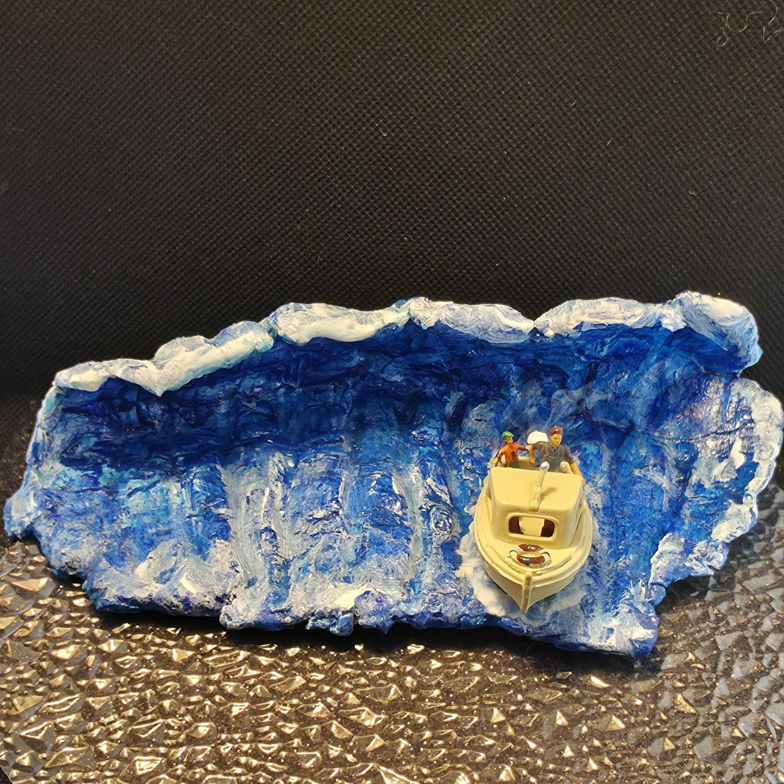 Sculpture en argile peinte représentant une vague tsunami ou raz-de-marée déferlante qu'essaie de fuir un petit bateau à moteur