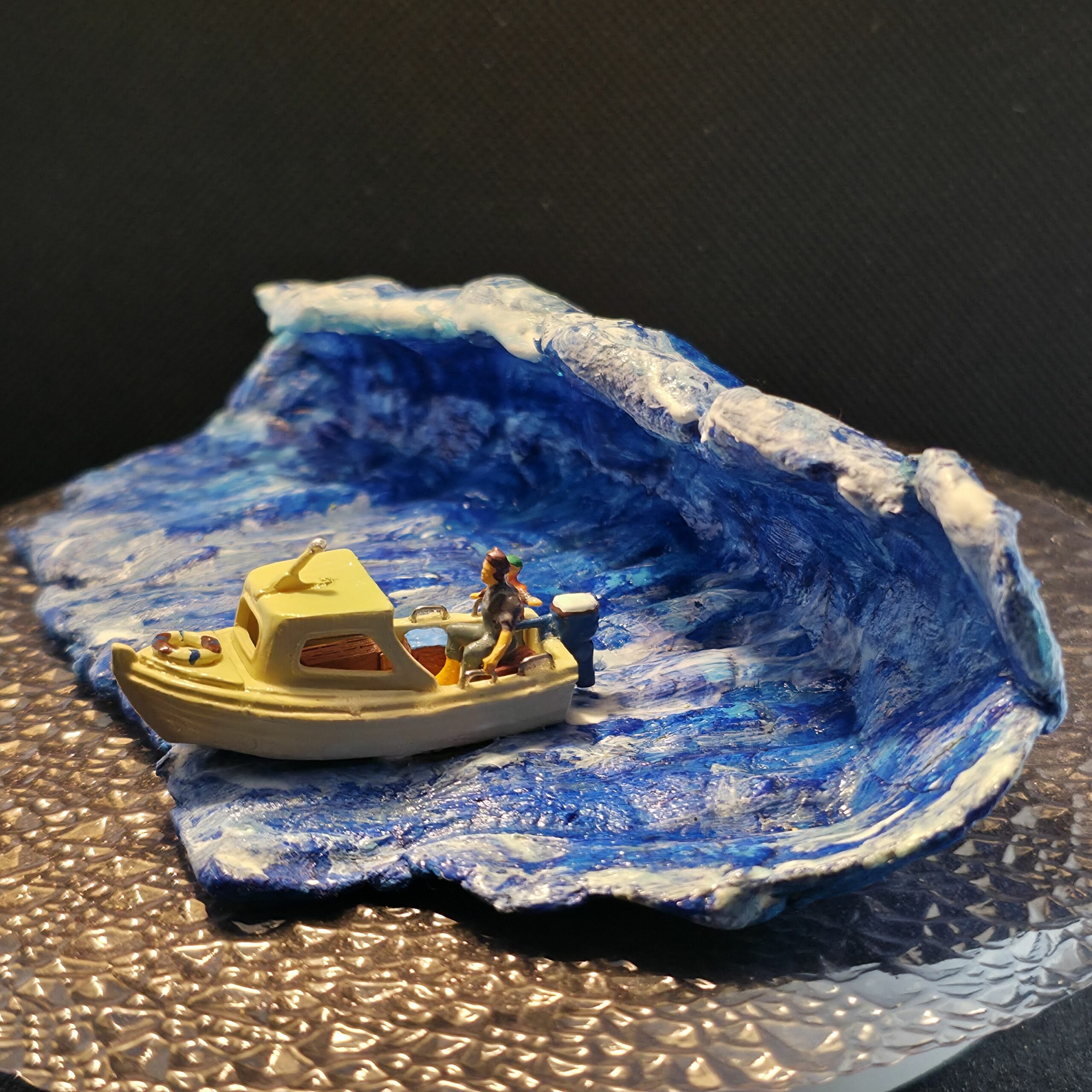 Sculpture en argile peinte représentant une vague tsunami ou raz-de-marée déferlante qu'essaie de fuir un petit bateau à moteur